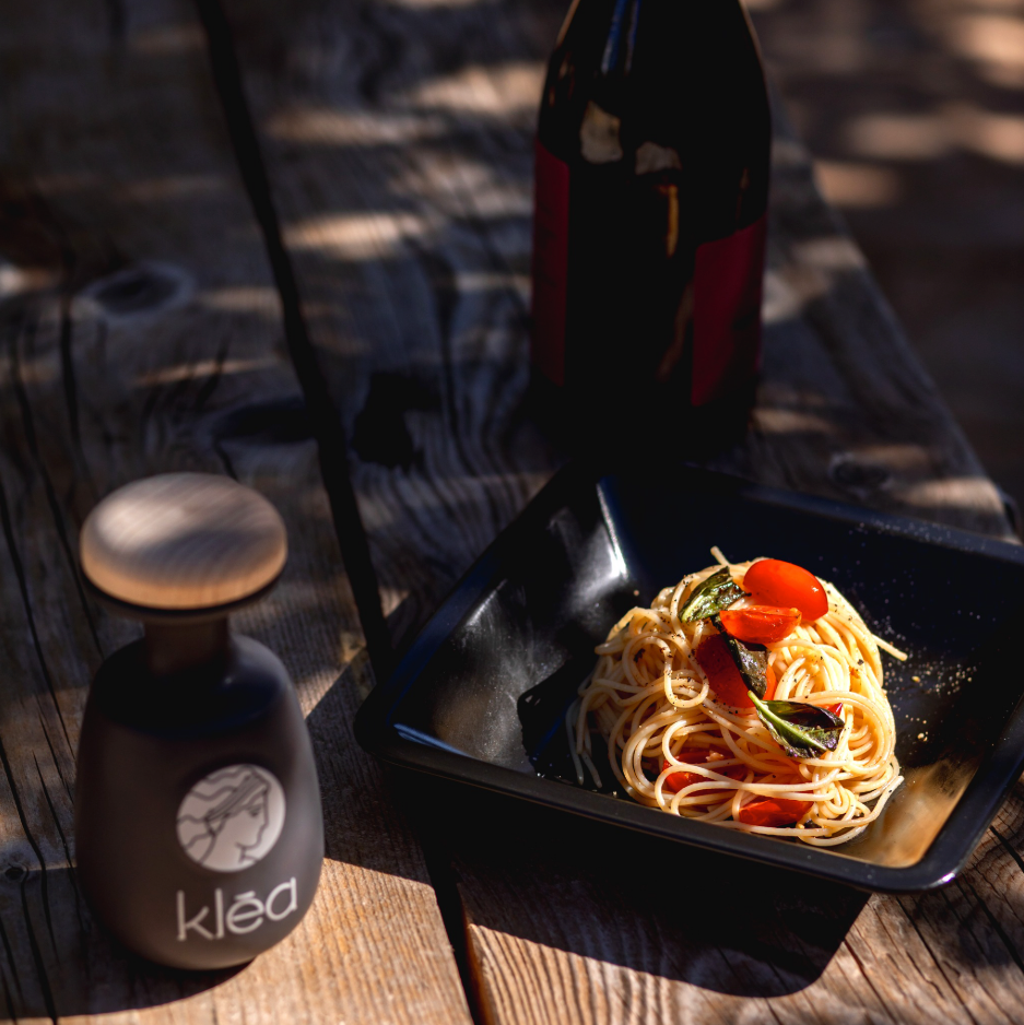 Klea Koroneiki Olivenöl in schwarzer Keramikflasche neben Pasta mit Tomaten und Kräutern angerichtet – natives Olivenöl extra Griechenland