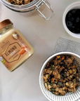 Mandelcreme ohne Zuckerzusatz glutenfrei vegan und ohne Palmöl im Glas neben Frühstücksschale mit Granola und Blaubeeren auf hellem Tisch